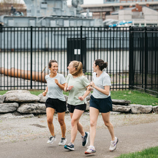 Three women running along a path by a waterfront with a black metal fence and buildings in the background.