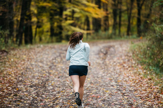 A woman running on a wooded path on a Fall day in her Middle Trail Running blue shorts and blue quarter zip.
