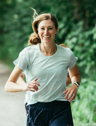 A women running, smiling in the blue t-shirt as she runs down a gravel path.