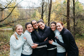 A group of 6 women all hugging each other on a Fall day wearing Middle Trail Running quarter zips to keep warm. 