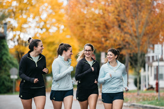 A group of four woman all running close to one another smiling on a fall day wearing their Middle Trail shorts and quarter-zip. 