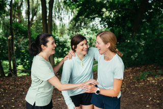 Three women standing together, linking arms, and laughing in their Middle Trail women's running apparel. 