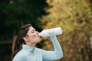 A woman drinking out of a white water bottle that has the light blue Middle Trail sticker with the Middle Trail logo. 