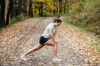 A women stretching before she begins her run on a Fall day as she wears her Middle Trail Running shorts and sage green t-shirt.
