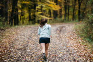 A woman running on a wooded path on a Fall day in her Middle Trail Running blue shorts and blue quarter zip. 