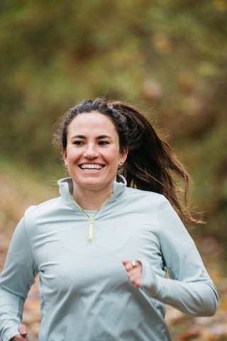 A close-up of a woman runner mid-stride, smiling in her light blue Middle Trail quarter zip.