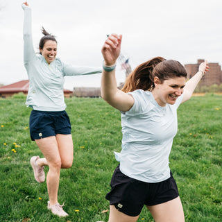 two women running down a hill, one in light blue running tee and black running shorts, and other in light blue running quarter zip and blue running shorts