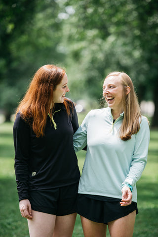 Co-Founders Genny and Kayla standing next to each other, linking arms and laughing, in their Middle Trail Running shorts and quarter zip.