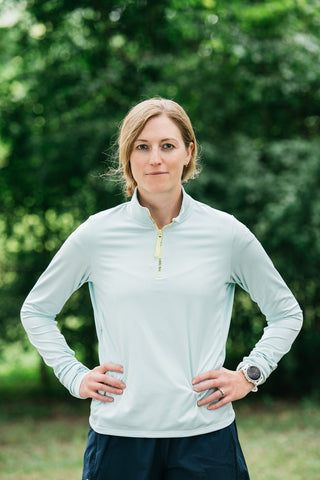 A women standing with her hands on her hips wearing the light blue Middle Trail women's running quarter zip.