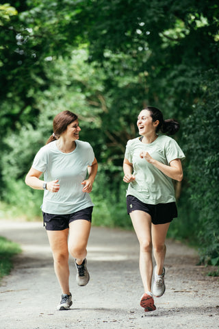 Women's Running Apparel. Two women running together in the Middle Trail running tee shirt and shorts on a wooded path. 