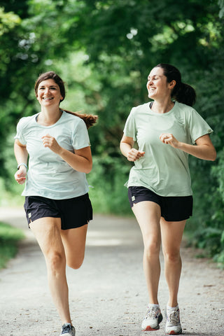 Two women running together laughing and wearing the black Middle Trail women's running shorts on a gravel path.