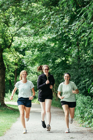 Three women running down a gravel path together in a row, with the woman in the middle wearing the black quarter-zip.