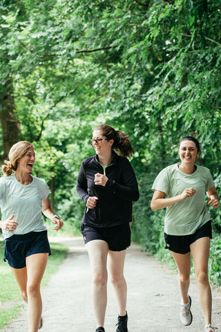 Three women running down a gravel path, laughing, with one woman wearing the blue shorts and the other two wearing black shorts.