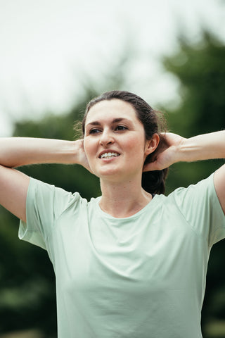 A women with the sage green running t-shirt on with her arms on her heads, recovering after a run. 
