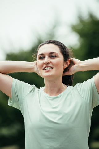 A woman with her hands on top of her head after a run wearing the sage green Middle Trail Running Breeze Tee-Shirt.