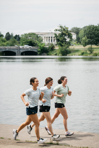 Three women running next to each other in sync while they run by Hoyt Lake in Buffalo, NY in Middle Trail Running women's apparel. 