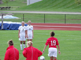 Co-Founders Genny and Kayla playing soccer in college. They are standing next to each other in a wall as they defend a free-kick in the soccer game.